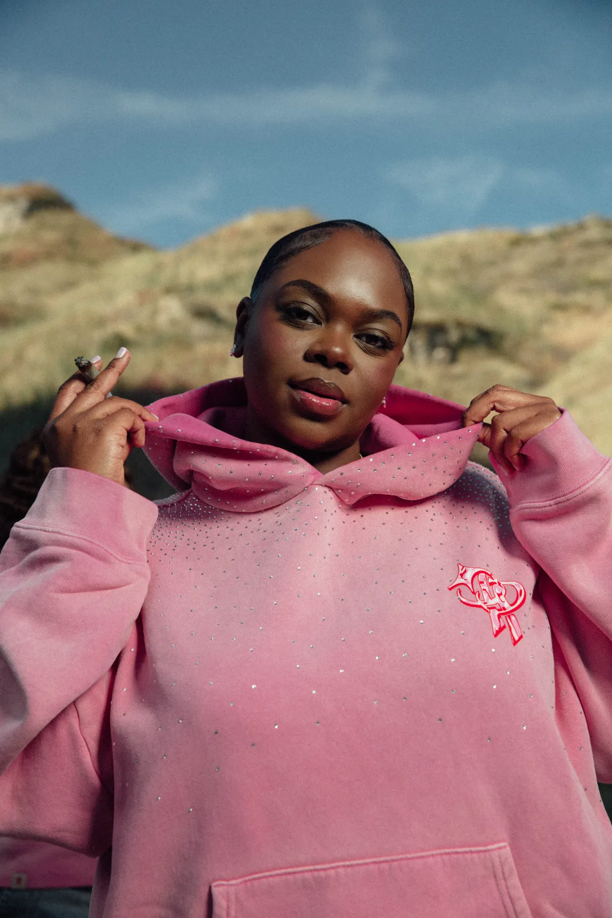 Campaign photo — model in the pink hoodie holding the hood open, sunlit hillside behind