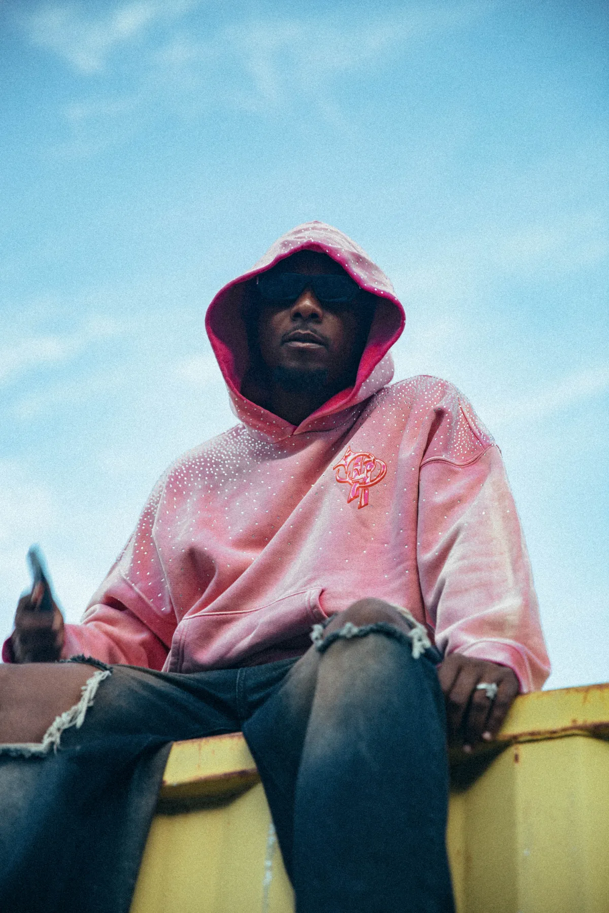 Campaign photo — model in the pink hoodie with the hood up, seated against open sky