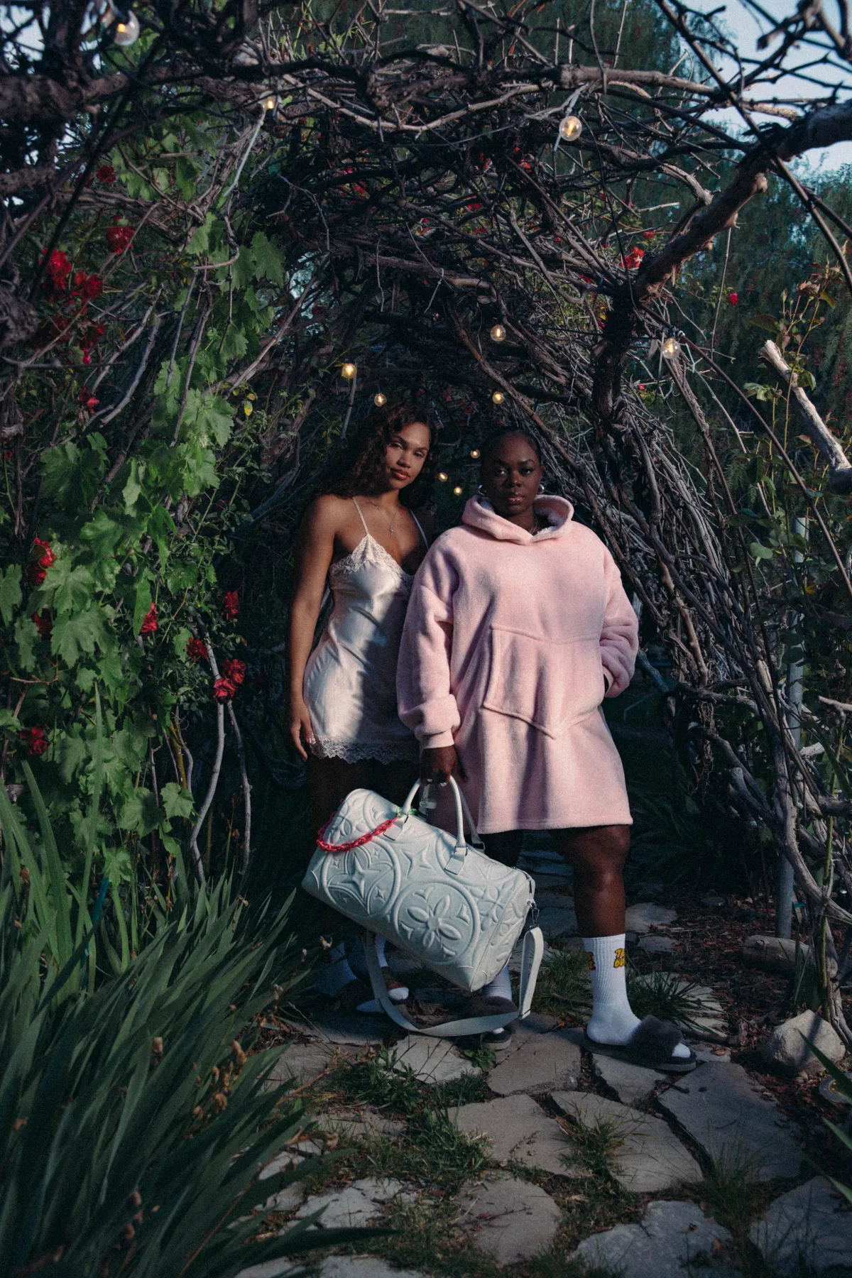 Campaign photo — two models under a garden archway at dusk, one carrying the Marshmallow Duffle